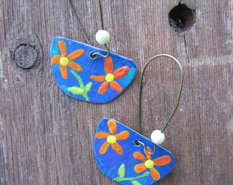 May include: Handmade earrings with a blue, semi-circular design, adorned with orange and yellow flower motifs. They have wire hooks and a small white bead. The background is a wooden surface.