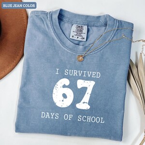 May include: A blue jean colored t-shirt with the text "I SURVIVED 67 DAYS OF SCHOOL" in white. The shirt has a crew neck and short sleeves. A gold necklace with a pendant is draped across the shirt.