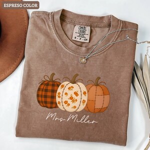 Pode incluir: Camiseta cor espresso com três abóboras ilustradas em cores de outono. As abóboras têm desenhos xadrez, florais e patchwork. O texto "Mrs. Miller" está escrito em itálico abaixo. Um colar de prata com um pingente está sobre a camiseta.