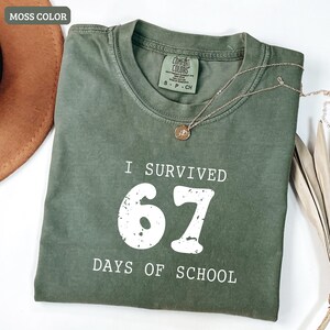 May include: A moss-colored t-shirt with the text "I SURVIVED 67 DAYS OF SCHOOL" in white. The shirt has a crew neck and short sleeves. A gold necklace with a pendant is draped on the shirt.
