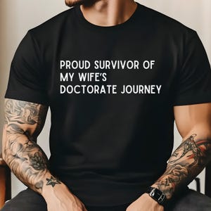 May include: A black t-shirt with white text that reads "Proud Survivor of My Wife's Doctorate Journey".