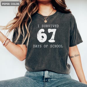 May include: Dark gray t-shirt with the text "I SURVIVED 67 DAYS OF SCHOOL" in white. The number 67 is large and distressed. The shirt is a crew neck and appears to be made of a soft material.