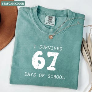 May include: Seafoam green t-shirt with the text "I SURVIVED 67 DAYS OF SCHOOL" in white. The shirt has a crew neck and short sleeves. A gold necklace with a pendant is draped across the shirt.