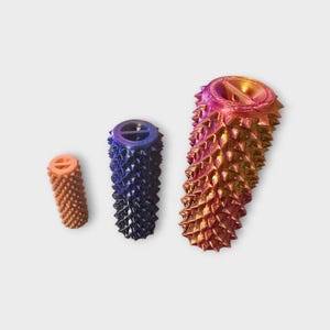 Könnte beinhalten: Drei 3D-gedruckte Objekte mit strukturierten, stacheligen Oberflächen. Das kleinste ist orange, das mittlere ist dunkelblau mit lila Akzenten und das größte ist ein Farbverlauf aus Pink, Orange und Gold. Jedes hat eine kreisförmige Oberseite mit einem Design.