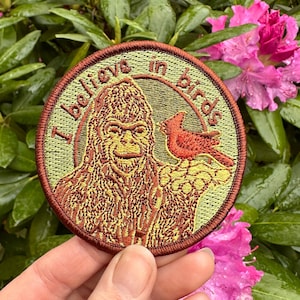 Peut inclure: Un écusson brodé marron et vert avec un Bigfoot et un oiseau rouge. L'écusson dit "Je crois aux oiseaux".