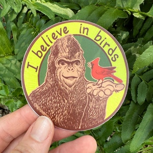 May include: A round sticker with a yellow background and green circle in the center. The text "I believe in birds" is written in brown around the green circle. A brown Bigfoot is holding out a red cardinal bird.