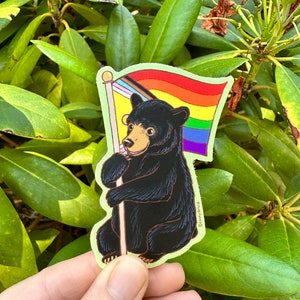 May include: A black bear sticker holding a rainbow pride flag. The flag has the colors red, orange, yellow, green, blue, indigo, and violet. The sticker is on a green leafy background.