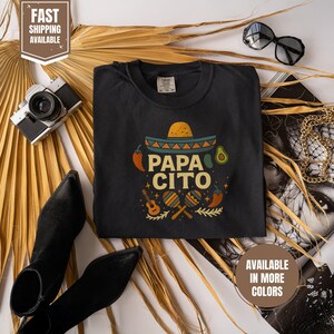 以下が含まれることがあります： ソンブレロ、アボカド、唐辛子、マラカスが描かれた、カラフルな「Papa Cito」グラフィックの黒いTシャツ。 サングラス、カメラ、ブーツなどのアクセサリーがシャツの周りにあります。