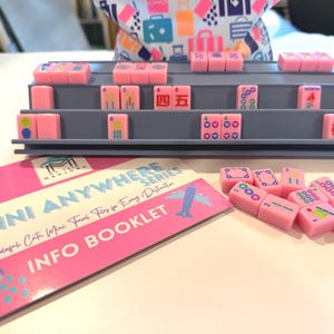May include: A pink Mahjong set with gray shelving and a booklet. The tiles are pink with various symbols and numbers. The booklet has pink and white text that reads "THE MINI ANYWHERE SERIES INFO BOOKLET."
