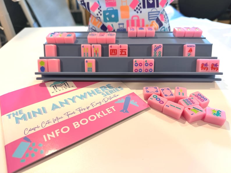 May include: A pink Mahjong set with gray shelving and a booklet. The tiles are pink with various symbols and numbers. The booklet has pink and white text that reads "THE MINI ANYWHERE SERIES INFO BOOKLET."