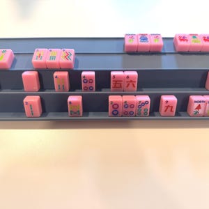 May include: A display of pink Mahjong tiles arranged on a gray tiered shelf. The tiles feature various symbols and numbers in different colors. The shelf is on a white surface.