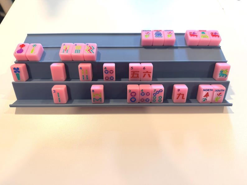May include: A display of pink Mahjong tiles arranged on a gray tiered shelf. The tiles feature various symbols and numbers in different colors. The shelf is on a white surface.