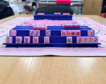 Puede incluir: Un soporte de plástico azul para fichas de Mahjong con fichas rosas que muestran varios símbolos, números y texto direccional, incluyendo North, South y West. El soporte está sobre una alfombrilla rosa claro con estampado floral y borde geométrico.