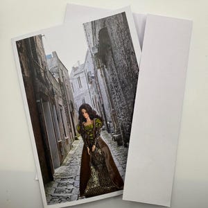 Könnte beinhalten: Ein Foto einer Puppe in einem langen Kleid im mittelalterlichen Stil, die in einer engen Kopfsteinpflasterstraße steht. Die Puppe hat lange braune Haare und trägt ein grünes und goldenes Oberteil mit einem braunen Rock und Mantel. Der Hintergrund zeigt alte Steingebäude.