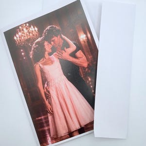 May include: A rectangular greeting card featuring a printed photograph of two people dancing in a ballroom. One person wears a light pink sleeveless dress with a full skirt, while the other wears a black shirt. A plain white envelope is placed beside it.