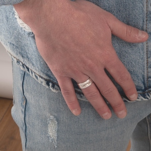 Könnte beinhalten: Ein silberner Ring mit hebräischen Schriftzeichen wird gezeigt. Der Ring befindet sich an einem Finger, wobei der Text deutlich sichtbar ist. Der Ring ist vor dem Hintergrund einer Jeansjacke und Jeans abgebildet.