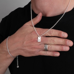May include: A silver necklace with a rectangular pendant engraved with the letter "R". A matching silver bracelet and a silver ring are also visible. The jewelry is displayed against a black background.