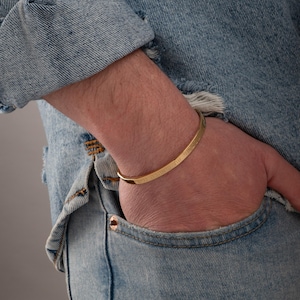 Puede incluir: Una pulsera dorada con texto grabado. La pulsera es curva y se lleva en la muñeca. La persona lleva una chaqueta vaquera y vaqueros. La pulsera es un accesorio sencillo y elegante.