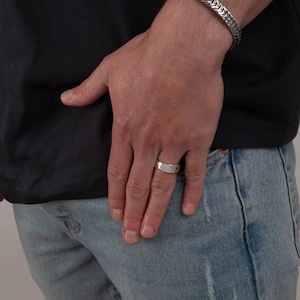 May include: A silver ring with a rectangular design is worn on a finger. A silver bracelet with a chain design is on the wrist. The person is wearing a black t-shirt and blue jeans.