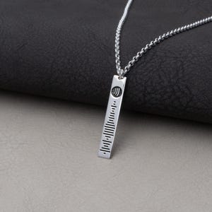 May include: A silver pendant necklace with a rectangular bar pendant. The pendant has a Spotify code design and a small Spotify logo at the top. The necklace chain is also silver and has a link design. The background is a dark grey surface.