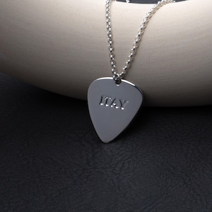 May include: A silver guitar pick pendant necklace. The pendant is shaped like a guitar pick and has the word "ITAY" engraved on it. The necklace chain is a delicate silver chain.