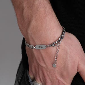 May include: A silver-colored bracelet with a curb chain design. The bracelet features a rectangular plate engraved with the name "Ron". The adjustable chain has a small circular charm at the end. The bracelet is worn on a wrist.