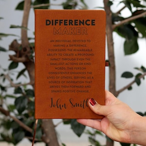 May include: A brown leather journal with the words "Difference Maker" embossed on the front. The journal has a quote about making a difference and a personalised name, "John Smith", at the bottom.