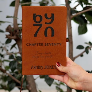 May include: A brown leather-bound journal with the text "by 70" and "Chapter Seventy" in black. The journal also includes the inscription "Every chapter brings new growth" and the name "Ashley JONES". A ribbon bookmark is visible.