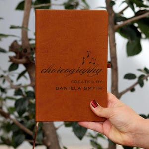 Könnte beinhalten: Ein braunes Leder-Notizbuch mit dem Wort "choreography" in eleganter Schrift, begleitet von Noten. Der Text "CREATED BY DANIELA SMITH" steht darunter. Ein braunes Lesezeichenband ist sichtbar.