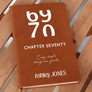 May include: A brown leather journal with the text "by 70" and "Chapter Seventy" in white. The journal also includes the quote "Every chapter brings new growth..." and the name "Ashley JONES". The journal is on a wooden surface.