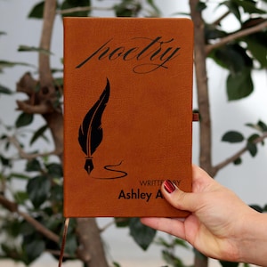 Pode incluir: Um diário de couro castanho com a palavra "poetry" em escrita cursiva e um desenho de pena preta. O diário está inscrito "WRITTEN BY Ashley A". Uma fita de marcador castanha é visível.
