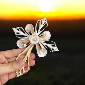 May include: A handcrafted flower made from seashells, featuring a central spiral and petal-like structures. The flower is held against a sunset backdrop, showcasing its natural colors of white, beige, and hints of yellow and brown. A unique coastal-themed decorative item.