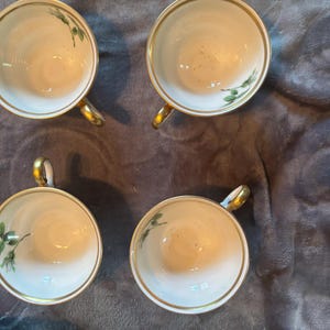 Può includere: Quattro tazze da tè bianche con bordi e manici dorati sono disposte a quadrato. Ogni tazza presenta un delicato motivo floreale verde. Le tazze sono appoggiate su una superficie scura e strutturata.