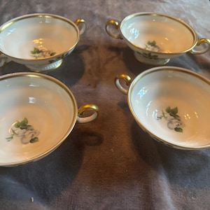 Könnte beinhalten: Vier weiße Porzellan-Suppenschalen mit goldenem Rand und Griffen. Jede Schale hat ein Blumendesign mit einer weißen Rose und grünen Blättern. Die Schalen sind auf einer neutralfarbenen Oberfläche angeordnet.