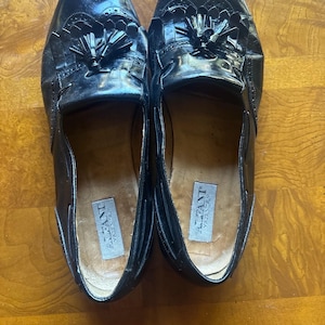 Puede incluir: Un par de mocasines de cuero negro con detalles de borlas. Los zapatos tienen un acabado brillante y un diseño clásico. El interior de cada zapato presenta una etiqueta con el texto "ALFANT VAZZA ITALY".