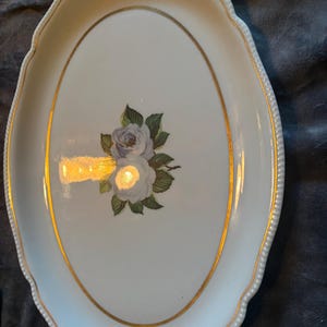 Könnte beinhalten: Ovale, cremefarbene Keramikservierplatte mit Goldrand und Blumendesign. Die Platte hat einen goldenen Rand und einen Perlenrand. Das Blumendesign zeigt zwei weiße und violette Rosen mit grünen Blättern.