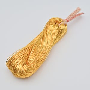 May include: A bundle of thin, metallic gold-colored threads or wires, neatly looped and gathered together. The strands are secured at one end with a small, light-colored braided cord, creating a long, slender, and flexible bunch of shiny material.