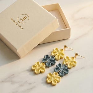 May include: Earrings with a floral design in yellow and blue-gray. The earrings are displayed next to an open beige box with the text "MINIMALIT BOX". The earrings have gold-colored posts and are on a white marble surface.