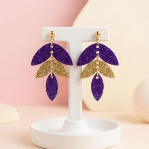 Peut inclure: Boucles d'oreilles en forme de feuilles pailletées violettes et dorées. Chaque boucle d'oreille est composée de trois feuilles superposées : violettes en haut et en bas, et dorées au milieu. Elles sont présentées sur un présentoir blanc.