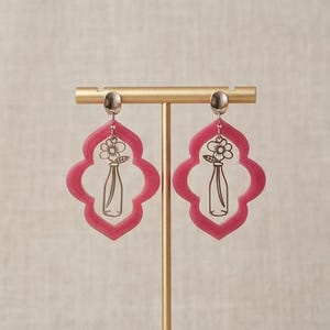 May include: A pair of dangle earrings featuring a quatrefoil shape in a translucent magenta hue. Each earring has a silver-colored top and a cutout of a vase with a flower inside. The earrings are displayed on a gold-colored stand.