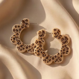 May include: A pair of cream-colored, flower-shaped hoop earrings with a leopard print pattern. The earrings are made of a smooth material and have a post closure. The earrings are displayed on a beige fabric.