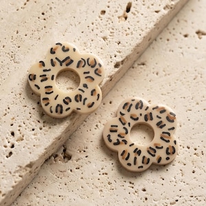 Peut inclure: Deux boucles d'oreilles en forme de fleur avec une base crème et un motif léopard. Les boucles d'oreilles ont un trou circulaire au centre et sont posées sur un fond neutre.