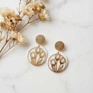 May include: Gold-colored tulip earrings with white circular backgrounds. The earrings have a textured gold stud and a gold tulip design. The earrings are displayed on a white marble surface with dried flowers.