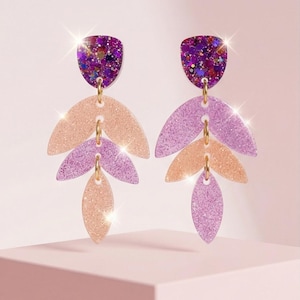 May include: A pair of dangling earrings with a layered leaf design. The top portion is a purple glittery shield shape. The leaves are in shades of peach and purple, connected by gold-colored hoops. The earrings are displayed on a pink surface.
