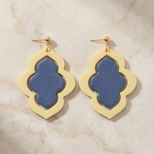 May include: A pair of cream and blue earrings. Each earring features a cream-colored, quatrefoil-shaped frame with a blue, textured center. The earrings have gold-colored posts and ball studs.