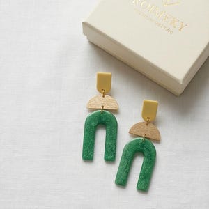 May include: A pair of geometric earrings with a green arch, a light wood half-circle, and a yellow rectangle. The earrings are displayed on a white surface next to a cream-colored box with gold text that reads "ROIMEKY PREMIUM GETTING".