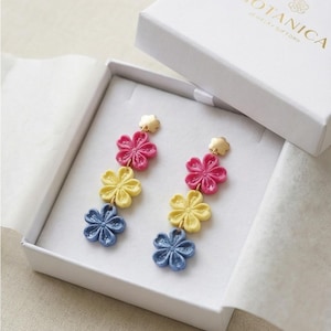 May include: A pair of floral earrings in a white gift box. The earrings feature three stacked flowers in pink, yellow, and blue. The box is labeled "BOTANICA JEWELRY GIFTS".