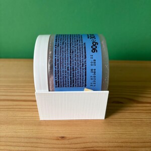 Könnte beinhalten: Ein durchsichtiger Kunststoffbehälter mit weißem Deckel, der Heidelbeer-Käsekuchen-Pulver enthält und in einem weißen, 3D-gedruckten Kunststoffständer steht. Das blaue Etikett zeigt Text wie 90g, 31.07.26, L2325064 28B sowie Zutaten in Deutsch und Englisch.