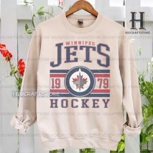 May include: Beige sweatshirt with the text "Winnipeg Jets" in blue and red, with "1979" and a maple leaf logo. The word "Hockey" is below the logo. The sweatshirt is suspended from a wooden hanger.