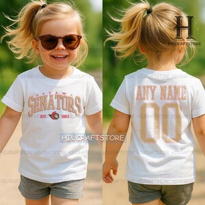 Puede incluir: Una camiseta blanca con el texto "OTTAWA SENATORS" en un estilo vintage, con el año "1883" debajo. La parte trasera de la camiseta tiene "ANY NAME" y el número "00". La camiseta la lleva un niño.